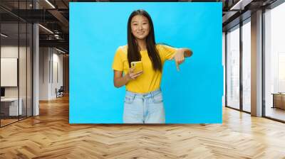 Happy Asian woman holding a phone with a yellow case on a blue background in a yellow T-shirt with a smile and teeth pointing her finger at Wall mural