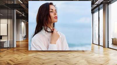 Portrait of a beautiful pensive woman with tanned skin in a white beach shirt with wet hair after swimming on the beach ocean sunset light with clouds Wall mural