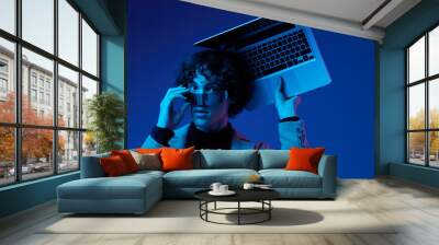 Portrait of a stylish male hacker with a laptop and futuristic glasses in blue light, cyber security, technology and education, neon color, freelancer online work Wall mural