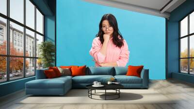 Sad cute Asian student young lady in pink hoodie sweatshirt wear round glasses recline on hand posing isolated on blue studio background. The best offer for ad. Eyewear for vision correction concept Wall mural