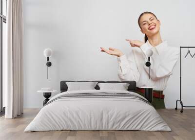 Smiling woman in a white shirt showing something with her hands, standing against a plain light background The concept conveys positivity and enthusiasm Wall mural