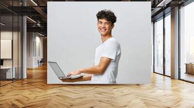 Smiling young man using a laptop with a neutral background, dressed casually in a white t shirt, suggesting positivity and modern technology engagement Wall mural