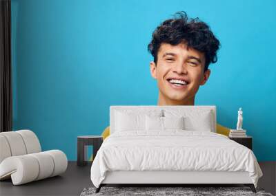 Smiling young man with curly hair in a bright yellow t shirt against a vibrant blue background, conveying happiness and energy, ideal for lifestyle and youth oriented concepts Wall mural