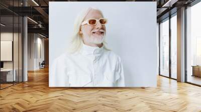 Stylish elderly man with long white hair and glasses, smiling against a light grey background, dressed in a modern white outfit, exuding confidence and charm Wall mural