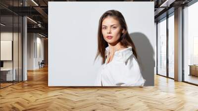 Woman with long hair in a white shirt, posing against a soft white background, showcasing natural beauty and elegance in a minimalist style Wall mural