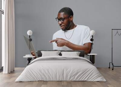 Young man using a laptop, wearing glasses, casual white t shirt, displaying excitement, neutral gray background Modern technology, lifestyle, and engagement with digital devices Wall mural