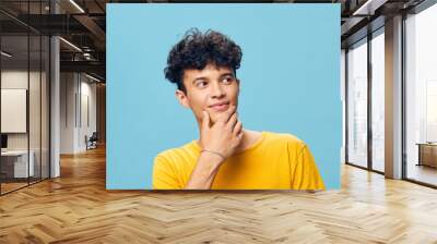 Young man with curly hair in a yellow shirt thinking thoughtfully on a blue background, expressing curiosity and contemplation in a cheerful manner Wall mural