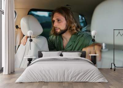 Young man with long hair sitting in a car, leaning forward and looking concerned or focused while wearing a green shirt Wall mural