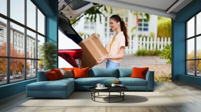 Young woman loading a cardboard box into her car, working outdoors She wears casual summer attire with a cheerful expression, surrounded by greenery and a cozy home backdrop Wall mural