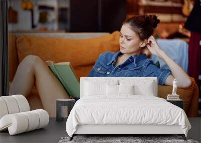 Young woman sitting on couch with crossed legs reading a book in denim shirt leisure activity at home Wall mural