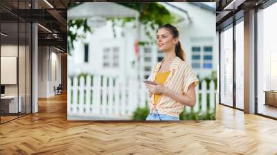 Young woman using smartphone outdoors, smiling joyfully in casual summer attire Bright colors enhance the cheerful vibe of a sunny day Green trees and a white house provide a fresh backdrop Wall mural