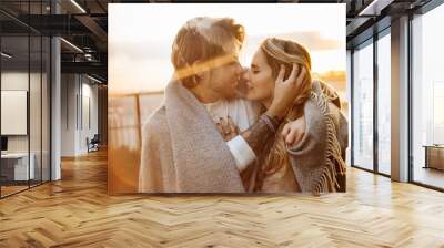 Portrait of happy beautiful couple at the sunset, cuddling in cozy blanket. Charming loving blond man hugging attractive woman, enjoying tenderness, spend time together, romantic date concept Wall mural