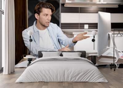 Serious male employee is sitting at the desk in home office, using computer, having a video conference. Focused young businessman talking with colleagues at online meeting, discussing a project Wall mural