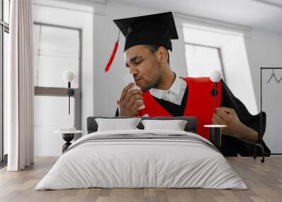 A Black student in graduate gown and square cap who is happy to finish his studies shows his the long-awaited diploma Wall mural