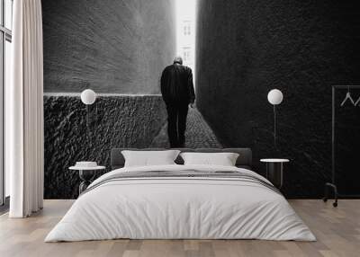 A man in black walking along a narrow alley toward the light. Black and white photography. Wall mural