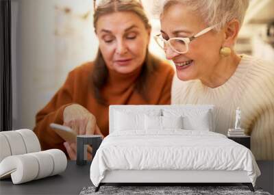 Aging, technology and communication concept. Cheerful two attractive mature female friends surfing internet together using generic cell phone. Elderly sisters watching funny videos online on mobile Wall mural