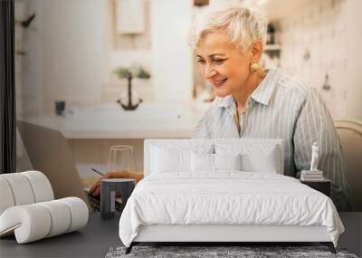 Attractive middle aged female freelancer in striped shirt sitting at home in front of open laptop, typing, working on content for website, using high speed internet connection, enjoying online work Wall mural