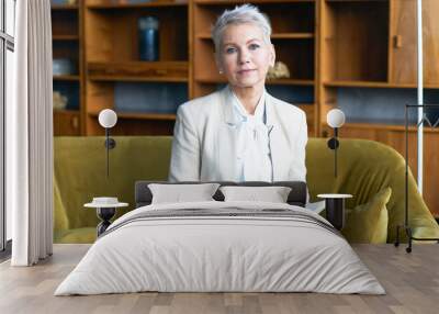 Attractive short haired middle aged female CEO sitting on comfortable couch in office interior writing in her notebook, checking appointment list, looking at camera with serious confident expression Wall mural