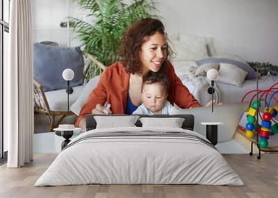 Attractive young dark skinned woman working at desk at home using laptop, holding baby on her lap. Portrait of smiling mother writing post on moms blog while her infant son playing with toy Wall mural