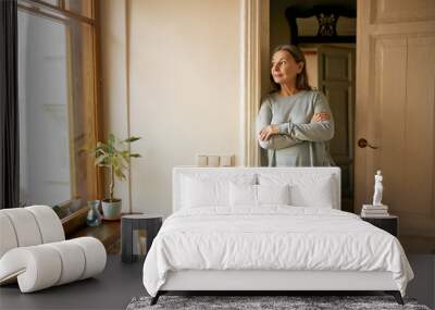 Beautiful casually dressed female pensioner posing indoors standing by windowsill with house plants, looking through window with pensive facial expression, leaning against white wall, daydreaming Wall mural