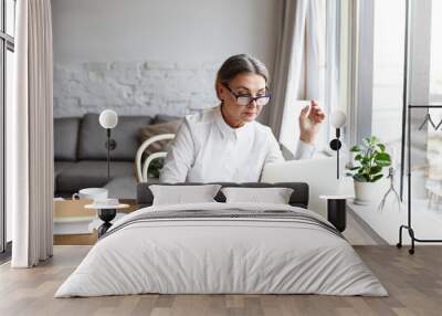 Beautiful concentrated middle aged businesswoman wearing rectangular glasses while keyboarding on generic laptop computer, sitting at her spacious office with notebook and cup of coffee on desk Wall mural