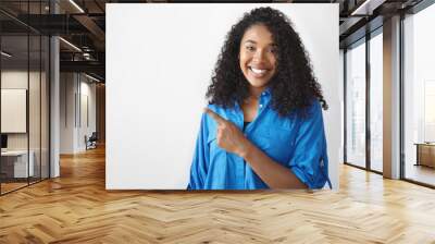 Beautiful young dark skinned woman dressed in blue shirt looking at camera with broad cheerful smile, pointing her index finger away at studio wall, demonstrating copy space for your content Wall mural