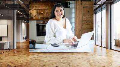 Beautiful young female accountant with curvy body and chubby cheeks typing on laptop, making calculations, providing outsource accounting service to various companies, working from home office Wall mural