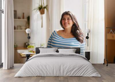 Beauty, bodypositivity, people and lifestyle concept. Indoor shot of gorgeous brunette Latin girl posing by window in kitchen dressed in xxl blue and white striped shirt, having joyful beaming smile Wall mural