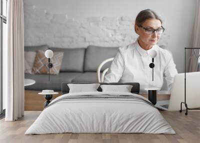 Busy modern mature female wearing spectacle and formal shirt working distantly using free wifi on generic laptop, sitting at wooden desk with books, having focused concentrated facial expression Wall mural