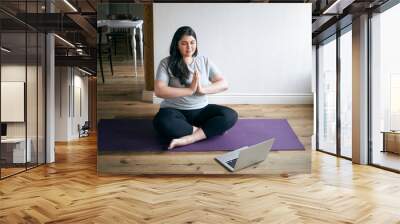 Calm mindful barefoot young female with curvy body practicing meditation at home, sitting in padmasana with eyes closed, holding hands pressed together in namaste, doing breathing exercises Wall mural