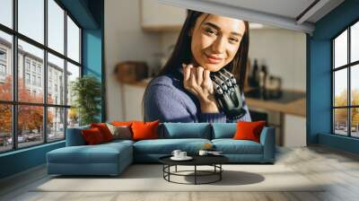 Close-up of charming lovely sweet young woman with sensual pouty lips and bionic black metal prosthesis instead of left arm looking at camera with happy sun drenched face, against kitchen background Wall mural