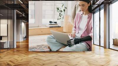 Close-up picture of beautiful charming female in pale pink silk shirt sitting on floor on colorful carpet holding laptop on knees with prosthetic bionic hand made of black metal mechanical device Wall mural