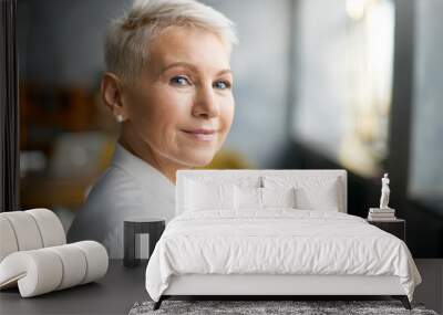 Close up portrait of attractive mature Caucasian woman with blonde pixie haircut and wrinkles looking at camera with confident smile while working in her office. Business, work, job and profession Wall mural