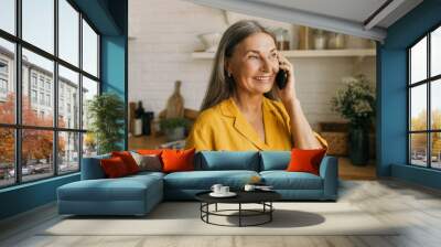 Close-up portrait of charming retired lady in yellow shirt having nice phone conversation isolated over kitchen interior background, looking through window during talk on black smartphone Wall mural