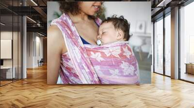 Close up shot of adorable cute newborn kid sucking on pacifier while sleeping peacefully in baby carrier at his mother's breast. Cropped image of young dark skinned mother carrying son in wrap Wall mural