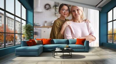 Cozy family portrait of stylish cute teenage girl in eyeglasses embracing her charming beautiful middle aged mother. Attractive neat blonde female posing in kitchen with her young daughter Wall mural