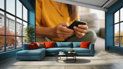 Crop shot of elderly woman's hands holding black mobile phone against background of her yellow blouse and green jeans, sitting on couch, spending leisure time in social networks, texting message Wall mural