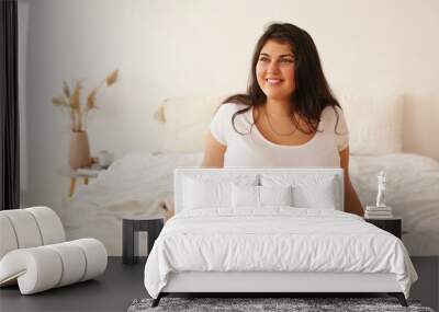 Cropped picture of positive cheerful young woman with chubby cheeks and long black hair sitting on edge of double bed having joyful happy facial expression, going to have walk right after awakening Wall mural