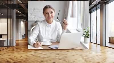 Elegant mature female psychologist working in modern office, holding eyeglasses and writing down in her diary, sitting at dews, using portable computer, smiling confidently, enjoying her work Wall mural