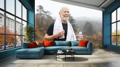 Exhausted elderly male with gray hair and beard drinking water after outdoor exercise and wiping sweat with towel around his neck. Tired but happy old senior man resting after running workout Wall mural