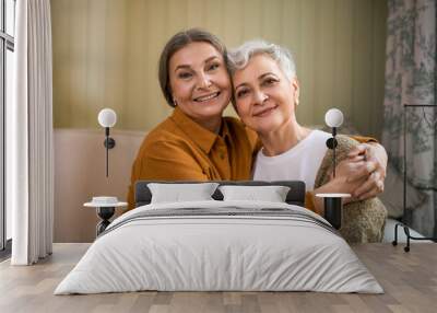 Family, age and mature people concept. Portrait of two joyful elderly sisters with wrinkles and gray hair sitting indoors on comfortable couch, embracing, happy to see each other after lock down Wall mural