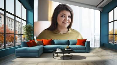 Friendly looking cheerful young lady with curvy body and loose dark hair enjoying beautiful urban view while standing by window at daylight, smiling happily at camera. People and lifestyle concept Wall mural