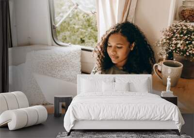 Gorgeous African girl with long black curly hair sitting near opened window at cozy bed with small fluffy pillows at traveling van with laptop on her knees, writing memories of her adventures on PC Wall mural