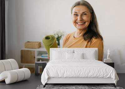 Happy cheerful joyful mature 60-years-old sporty woman standing against bed at her apartment with rolled green mat in hands, waiting with anticipation beginning of daily physical workout Wall mural