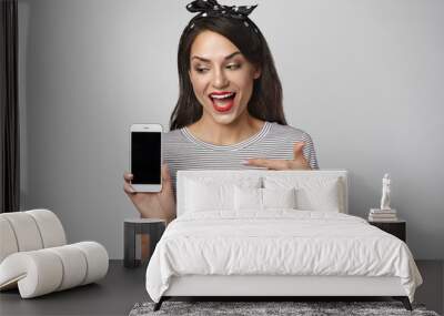 Happy emotional young female model with headband advertising gadgets in studio, opening mouth excitedly, pointing at white generic cell phone with copy space black screen for your information Wall mural