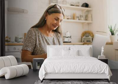 Happy senior woman making online payments of bill using laptop, smiling while shopping with credit card, using application for Internet banking, isolated against kitchen surroundings Wall mural