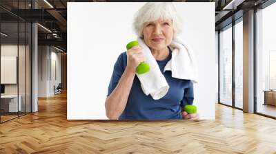 Health, wellness, activity, maturity and age concept. Upbeat elderly woman on retirement training indoors with white towel around her neck, exercising with pair of green dumbbells and smiling Wall mural