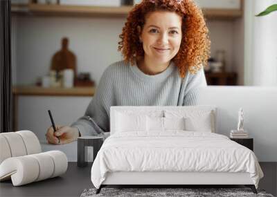 Home portrait of pretty female English tutor working at home sitting at kitchen table, preparing for online lesson with her student, writing in her copybook in front of laptop and cup of coffee Wall mural