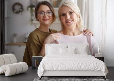 Horizontal image of cute young daughter in stylish hipster eyeglasses embracing her mother with love, spending day together, having happy joyful facial expressions, smiling broadly at camera Wall mural