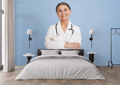 Horizontal image of happy successful mature gray haired female pediatrician posing at blue wall, keeping arms on her chest. People, age, healthcare jobs, occupations and medical careers concept Wall mural